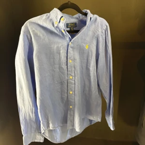 Blå skjorta från Polo Ralph Lauren - Snygg ljusblå skjorta från Polo Ralph Lauren med klassisk krage och knappar i gul ton. Ikoniska broderade loggan i gult på bröstet. Skjortan är långärmad och tillverkad i mjuk bomull, perfekt för en clean och stilren look.