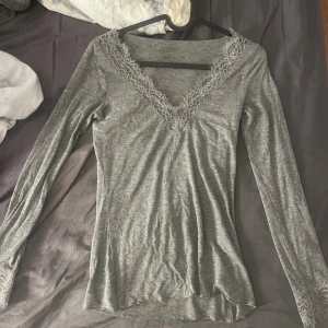 Intimissimi tröja grå - Snygg grå långärmad topp med v-ringning och spetsdetaljer vid halsringningen och ärmsluten. Tunn och mjuk i materialet, perfekt för dig som gillar stilrena plagg med lite extra detaljer. Passformen är normal och toppen har en feminin touch med spetsen.