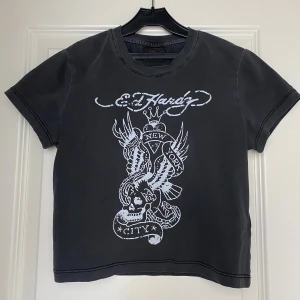 Svart croppad t-shirt Ed Hardy - Svart croppad t-shirt från Ed Hardy med vit NYC-grafik och döskalletryck på framsidan. Modellen är kort i midjan och har klassisk rund hals samt korta ärmar. Har endast provats.