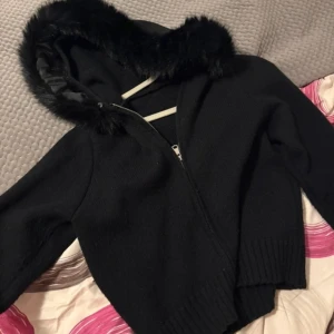 Svart cardigan med fuskpäls - Svart stickad cardigan med dragkedja och huva med fluffig fuskpäls.  Köpt för 800kr