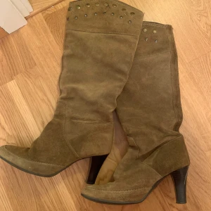 Bruna mockastövlar med klack och nitar - Snygga höga stövlar i brun/grön mocka med dekorativa nitar upptill. Väckar sig längst ner. Perfekt för dig som vill ha en cool och trendig stil med lite edge. Materialet är mjukt och ger en lyxig känsla.