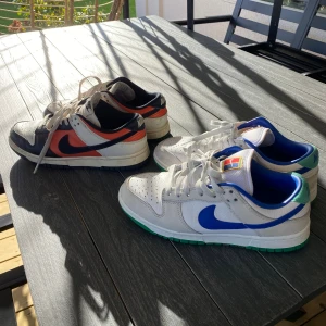Nike Dunk sneakers i två färger - Två par Nike Dunk sneakers säljes. 500kr st.  Ena paret är vitt, blått och grönt med blå swoosh och gröna detaljer på sulan. Det andra paret är vitt, svart och orange med svart swoosh. Båda har lågt skaft, snörning och platt sula. Perfekt för dig som gillar klassiska och färgstarka sneakers.