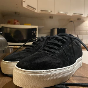 Svarta sneakers från ARIGATO i mocka - Snygga svarta sneakers från ARIGATO med ovandel i mjuk mocka och klassisk vit platt sula. Skorna har rund tå, svarta snören och diskret guldfärgad logga på sidan. Perfekt för dig som gillar stilrena och trendiga sneakers med premiumkänsla.