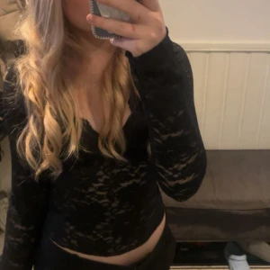 Svart spetsblus - Säljer en svart långärmad blus i genombruten spets med v-ringning. Toppen är croppad och har en figurnära passform som framhäver siluetten. Perfekt för dig som gillar en edgy och trendig look.