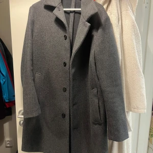 Grå rock från Zara i storlek L - Snygg grå rock från Zara i storlek L med klassisk krage och tre svarta knappar framtill. Jackan har två fickor på sidorna och är tillverkad i ett mjukt ullmaterial som känns lyxigt. Perfekt för dig som gillar stilrena och tidlösa plagg.