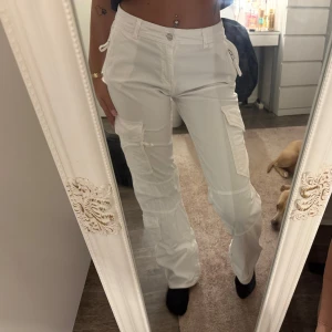 Vita cargopants med fickor - Säljer ett par vita cargopants med flera praktiska fickor på sidorna och bak. Byxorna har raka ben, midja med knapp och dragkedja samt coola detaljer som snörning vid fickorna. Perfekta för en avslappnad och trendig look.