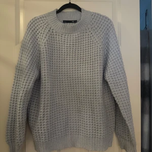 Ljusgrå stickad tröja från Gina Tricot - Supermysig ljusgrå stickad tröja med grovstickat mönster och rund hals. Tröjan har långa ärmar och breda ribbade muddar vid ärmslut och nederkant. Perfekt för kyliga dagar och enkel att styla till jeans eller kjol.