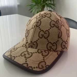 Gucci keps med GG-monogram - Snygg keps från Gucci i beige och brunt med det klassiska GG-monogrammet över hela kepsen. Justerbar rem baktill i brunt skinn och detaljerad med mörkbruna kanter. Tillverkad i bomullsmix och perfekt för dig som vill sticka ut med en lyxig accessoar.