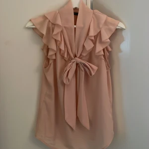 Ljusrosa volangblus från Morgan - Supersöt ljusrosa blus från Morgan med volangdetaljer vid axlarna och en stor knytrosett framtill. Ärmlös modell i ett lätt, flowy material som ger en romantisk vibe. Perfekt för dig som gillar feminina detaljer och vill sticka ut lite extra.