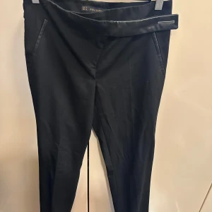 Svarta kostymbyxor från Zara Basic - Svarta kostymbyxor från Zara Basic med hög midja och snyggt omlottbälte med tryckknapp. Byxorna har smal passform, diskreta fickor och är tillverkade i ett mjukt tyg som känns skönt mot huden. Perfekta för en clean och stilren look.