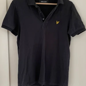 Svart pikétröja från Lyle & Scott S - Klassisk svart pikétröja från Lyle & Scott i storlek S som är använd ytterst lite. Tröjan har korta ärmar, krage och knäppning framtill samt den ikoniska gula fågel-loggan broderad på bröstet. Tillverkad i mjuk bomull för en skön känsla.