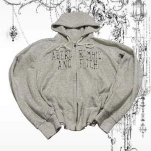 Grå hoodie från Abercrombie & Fitch - Säljer en ljusgrå hoodie från Abercrombie & Fitch med broderad logga framtill. Tröjan har dragkedja, huva med snörning och två fickor. Perfekt för chill dagar och enkel att matcha med jeans eller mjukisbyxor.