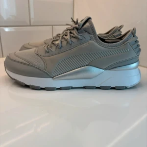 Gråa Puma RS-0 sneakers - Snygga Puma RS-0 sneakers i helgrått med silvriga detaljer och vit chunky sula. Skorna har mesh och syntet på ovandelen, samt snörning och coola detaljer på hälen. Perfekta för dig som gillar en modern och sportig look.
