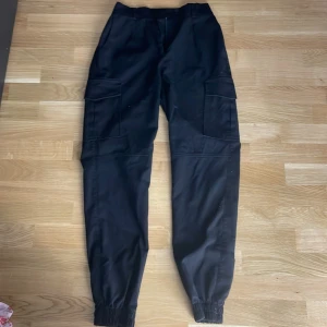 Svarta cargopants med resår, stl 34 - Svarta cargopants med stora fickor på sidorna och resår vid bensluten. Snygg och avslappnad passform, perfekta för en streetwear-look. Byxorna har klassisk cargomodell och är tillverkade i ett mjukt material som känns bekvämt hela dagen.