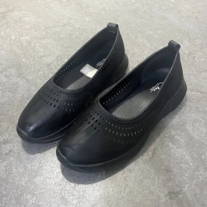 Svarta ballerinaskor i skinn från Clou - Svarta ballerinaskor från Clou i storlek 39. Skorna har rund tå, platt sula och är tillverkade i skinn med små perforerade hål för extra ventilation. Enkla att ta på med liten ögla bak. Perfekta för dig som gillar stilren och bekväm design.