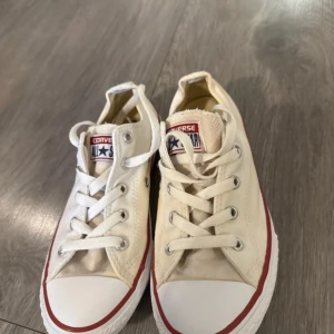 Vita Converse All Star sneakers - Klassiska vita Converse All Star sneakers i lågt snitt med röd rand längs sulan. Skorna har rund tå, platt sula och snörning framtill. Tillverkade i canvas med gummidetaljer och logga på plösen. Perfekta för en avslappnad och cool stil.