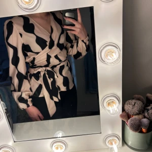 Beige och svart omlottblus från Shein - Säljer en beige och svart omlottblus från Shein med vågigt mönster. Blusen har långa ärmar med resår vid ärmslut, knytband i midjan och v-ringning. Tillverkad i polyester med en bekväm passform.