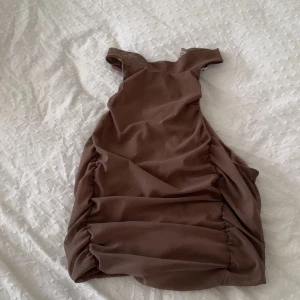  Brun omlotttopp  från NA-KD XS - Supersnygg brun kortklänning från NA-KD i omlottmodell med rynkade sidor. Klänningen är ärmlös och har en tight passform som framhäver figuren. Perfekt för dig som gillar trendiga och stilrena plagg. Materialet känns lätt och stretchigt.