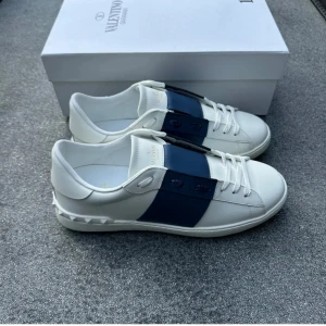 Valentino Garavani Open sneakers vit/blå - Snygga Valentino Garavani Open sneakers i vitt skinn med bred blå läderdetalj över mitten. Klassisk rund tå, vita snören och ikoniska nitar baktill på sulan. Perfekta för dig som vill ha en stilren och lyxig look med en twist.