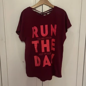 Vinröd t-shirt med tryck och korsad rygg - Vinröd t-shirt med stort rosa tryck 'RUN THE DAY' framtill och peppande text. T-shirten har korta ärmar och snygga korsade band i ryggen. Skön och luftig passform, perfekt för träning eller chill. Materialet känns mjukt och lätt.