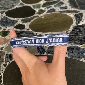 Blå Christian Dior J'Adior armband - Snyggt blått armband från Christian Dior med texten 'J'Adior' broderad i vitt. Armbandet har en cool boho-vibe med tofsar i ändarna och är gjort i ett mjukt tygmaterial. Perfekt för att ge din outfit en lyxig touch och visa din Dior-kärlek.