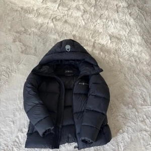 Svart dunjacka med huva från Sail Racing - Säljer en svart pufferjacka från Sail Racing med huva och dragkedja framtill. Jackan har en stilren design med logga på bröstet och är perfekt för kyliga dagar. Fylld med dun för extra värme och komfort. Säljes i nyskick använd endast fåtal gånger, säljer på grund av att den ej kommer till användning eftersom jag har en annan. Storlek medium 3000kr