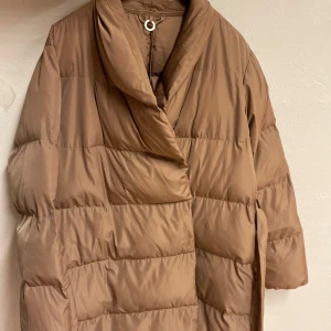 Beige pufferjacka med bälte - Säljer en beige pufferjacka från Stockh  LM med bred krage och omlottstängning. Jackan har ett matchande bälte i midjan och tryckknappar på insidan. Perfekt för kalla dagar med sin quiltade design och långa ärmar.