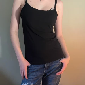 Svart linnetopp med smala axelband - En svart, enkel linnetopp med smala justerbara axelband och rak skärning. Toppen är figurnära och tillverkad i mjukt bomullsmaterial. Perfekt att styla med jeans eller kjol för en clean look.