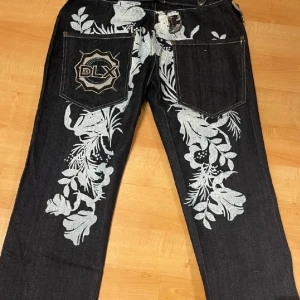 Deeluxe Wear jeans med blommigt tryck - Mörkblå jeans från Deeluxe Wear med stora bakfickor, broderad DLX-logga och unikt blommigt mönster i vitt på baksidan. Byxorna har loose fit, raka ben och är tillverkade i kraftig denim. Snygga kontrastsömmar och coola detaljer gör dem till ett statement-plagg.