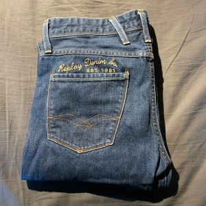 Replay Anbass jeans mörkblå herr - Snygga Replay Anbass jeans i mörkblå tvätt med klassisk femficksdesign och gul kontrastsöm. Bakfickorna har riktigt unika, broderade detaljer. Jeansen har rak passform och är tillverkade i slitstarkt denim.