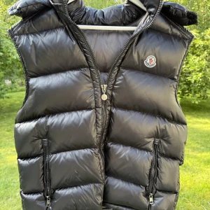Moncler väst - Snygg svart dunväst från Moncler med glansig finish och klassisk logga på bröstet. Västen har huva, två dragkedjefickor och quiltad design som ger en cool, puffad look. De finns 2 deffekter därav priset: deffekt 1 de är en tråd som har släppt men inget hål eller så och ett pyttelitet hål i insidan av fickan, Skriv om ni har några frågor eller funderingar 