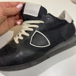 Svarta sneakers från Philippe Model - Snygga svarta sneakers från Philippe Model med vita detaljer och snörning. Skorna har en mix av skinn och textil, samt en ikonisk sköldlogga på sidan. Vit hälkappa och platt sula ger en clean look. Perfekta för dig som gillar stilrena och trendiga sneakers.