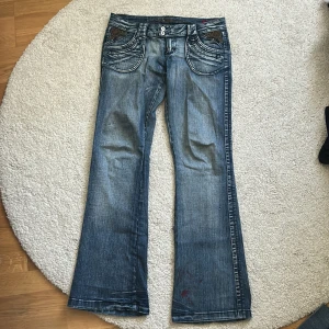 Blå bootcut jeans från Foxy Jeans - Säljer ett par blå bootcut jeans från Foxy Jeans med låg midja och snygga broderade detaljer på bakfickorna. Jeansen har markerade sömmar, dubbla knappar i midjan och stora fickor fram och bak. Perfekta för dig som gillar en retro vibe och unika detaljer.