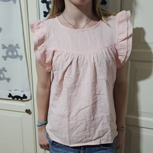 Ljusrosa blus med volangärmar - Supersöt ljusrosa blus i bomull med diskret strukturmönster och volangärmar. Rund halsringning och knapp i nacken. Luftig och lätt modell som ger en romantisk vibe. Perfekt till jeans eller kjol för en fräsch look. Aldrig använd eller tvättad, endast testad. Storlek 170