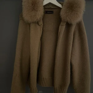 Beige stickad hoodie med pälskant - Mysig beige stickad hoodie från Alessa med fluffig pälskant runt huvan. Tröjan har långa ärmar, ribbade muddar och en relaxed passform. Perfekt för kyliga dagar när du vill vara både varm och snygg.