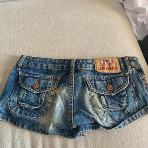Blå jeansshorts från Y.L.S Jeans - Snygga blå jeansshorts från Y.L.S Jeans med slitna detaljer och stora fickor bak med knappar. Klassisk femficksmodell, låg midja och cool tvättad look. Perfekta för sommaren och ger en avslappnad vibe.