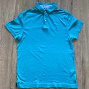 Turkos pikétröja från Tommy Hilfiger - Snygg turkos pikétröja från Tommy Hilfiger med klassisk krage och två knappar framtill. Liten broderad logga på bröstet. Tillverkad i mjuk bomull och har korta ärmar. Perfekt för en fräsch och stilren look.
