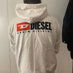 Vit hoodie från Diesel med dragkedja - Vit hoodie från Diesel med stor logga och texten 'Denim Division' på ryggen samt mindre logga på bröstet. Tröjan har huva med snörning, dragkedja framtill och två fickor. Nypris 1200kr