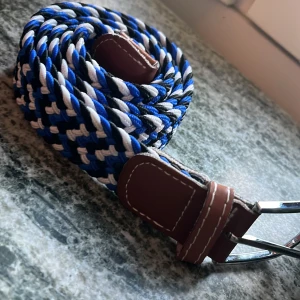 Randigt flätat bälte i blått och vitt - Snyggt flätat bälte med randigt mönster i blått, vitt och svart. Bältet har detaljer i brunt skinn och ett silvrigt spänne. Perfekt accessoar för att lyfta din outfit med lite färg och stil.