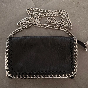 Svart axelväska med kedja från Tiamo - Cool svart axelväska från Tiamo i skinnimitation med grov silverfärgad kedja som axelrem. Väskan har dekorativa detaljer runt kanterna och stängs med dragkedja och magnetknappar. Perfekt storlek för det viktigaste och ger en edgy vibe till din outfit.