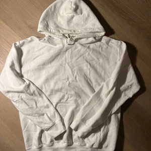 Vit hoodie med röd joker-tryck - Vit hoodie med huva och känguruficka. På ryggen finns ett stort rött tryck med en jokerfigur och texten 'JOKER' längs sidorna. Tröjan har långa ärmar och är gjord i mjuk bomull, perfekt för en chill och avslappnad stil.