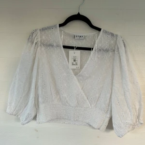 Vit broderad blus från Stay - Superfin vit blus från Stay med broderade blommor och omlott-v-ringning. Blusen har trekvartsärmar med resår vid ärmslut och smockad midja för en snygg passform. Perfekt till jeans eller kjol för en fräsch look.🤩💕