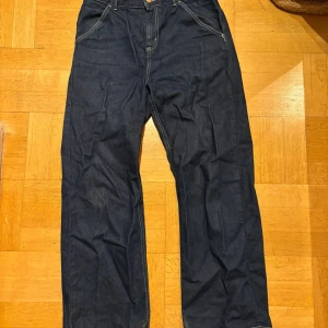 Carhartt WIP Simple Pant Loose Jeans - Mörkblå jeans från Carhartt Work In Progress, modell W' Simple Pant med loose fit. Klassisk femficksdesign, kontrastsömmar och Carhartt-logga på bakfickan. Tillverkade i 100% bomull för en robust känsla och avslappnad stil.