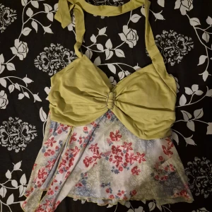 Blommig grön och vit linnetopp - Säljer en unik linnetopp med ljusgrönt överdel och knytband i nacken. Toppen har en snygg rosett-detalj framtill och en flowy, blommig nederdel i vitt med röda och blå blommor. Perfekt för dig som vill sticka ut med en färgglad och somrig look!