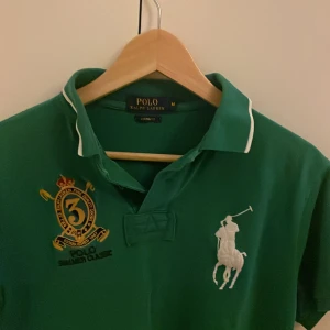 Grön Polo Ralph Lauren t-shirt M - Snygg grön t-shirt från Polo Ralph Lauren i custom fit med klassisk krage och vit kant. Stora broderade loggor på bröstet, inklusive polospelare och emblem med siffran 3. Perfekt för dig som gillar sportig och preppy stil.
