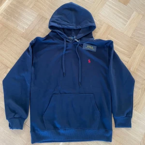 Marinblå hoodie från Polo Ralph Lauren - Snygg marinblå hoodie från Polo Ralph Lauren med klassisk huva och dragsko. Tröjan har en röd broderad logga på bröstet och en stor magficka framtill. Tillverkad i mjuk bomull och har ribbade muddar vid ärmslut och nederkant.