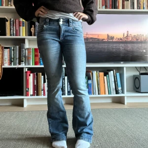 Blå bootcut lågmidjade jeans - Säljer ett par blå bootcut jeans med snygg tvätt och dubbla knappar i midjan. Jeansen har klassiska fickor fram och bak, samt en liten ficka med lock och knapp bak. Perfekta för dig som gillar en retro vibe och vill ha ett par jeans med lite utsvängda ben.