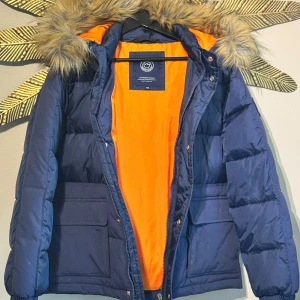 Oanvänd Short padded Parka med fuskpäls från Svea - Mörkblå dunjacka från Svea med orange foder, stor huva med fuskpäls och rejäla fickor framtill. Jackan har dragkedja och knappar, samt justerbar dragsko nertill. Perfekt för kalla dagar och riktigt snygg streetstil. helt oanvänd och har bara hängt i skåpet sedan inköp. 