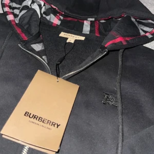 Svart Burberry hoodie med dragkedja - Svart hoodie från Burberry med klassiskt rutigt foder i huvan och broderad logga på bröstet. Tröjan har dragkedja framtill, snörning i huvan och ribbade muddar. Perfekt för dig som vill ha en stilren och lyxig look.