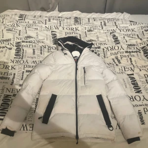 Vit pufferjacka från Zavetti Canada XS - Säljer en snygg vit pufferjacka från Zavetti Canada i storlek XS. Jackan har svarta detaljer, dragkedjor, huva och en patch på ärmen. Perfekt för kalla dagar med sin vadderade design och coola look. Använt skick och otvättad men annars inga defekter!